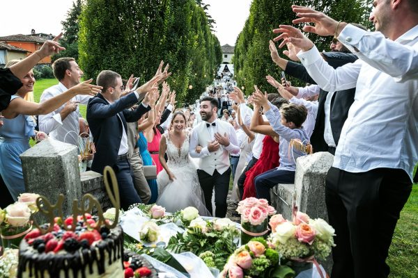 A newlywed couple walks through a cheering crowd of guests outdoors, with raised hands forming a celebratory aisle. Flowers and a decorated cake fill the foreground.