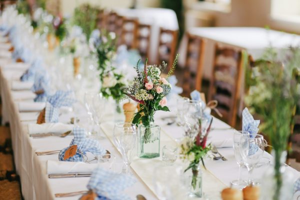 A long, elegantly set dining table at an indoor event, decorated with small floral arrangements in glass bottles, white tablecloths, clear glassware, silverware, and blue-and-white gingham napkins. Wooden chairs and additional decorated tables are softly blurred in the background.