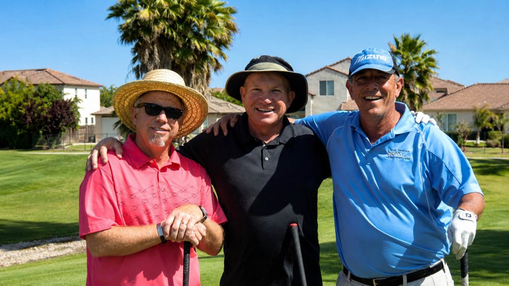 Three smiling golfers stand together on a sunny golf course with their arms around one another, wearing pink, black, and blue shirts and holding golf clubs.