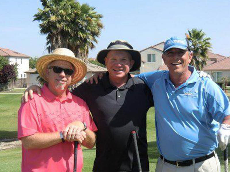 Three smiling golfers stand together on a sunny golf course with their arms around one another, wearing pink, black, and blue shirts and holding golf clubs.