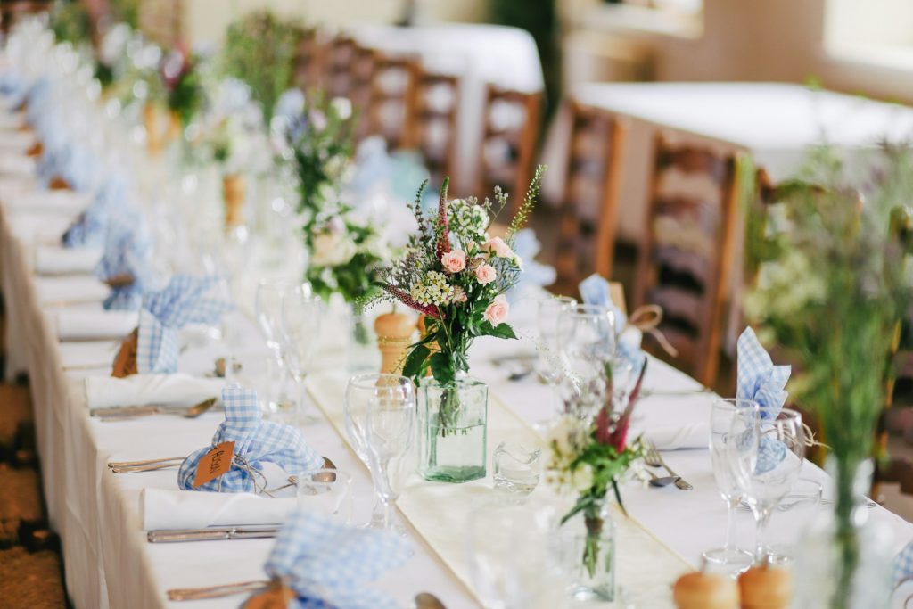 A long, elegantly set dining table at an indoor event, decorated with small floral arrangements in glass bottles, white tablecloths, clear glassware, silverware, and blue-and-white gingham napkins. Wooden chairs and additional decorated tables are softly blurred in the background.
